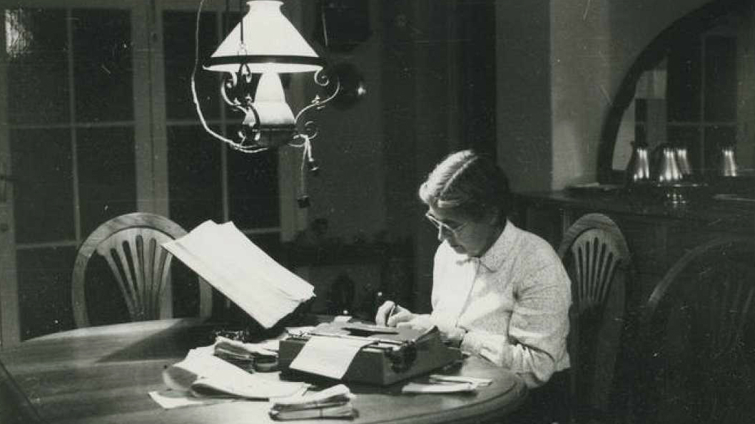 María Moliner escribiendo a máquina en la mesa de su casa, de noche, rodeada de papeles y fichas